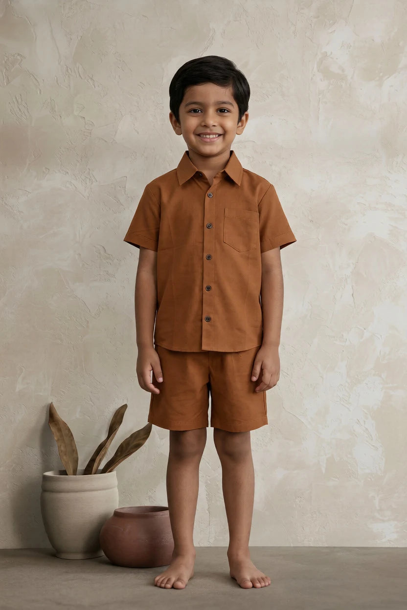Child wearing a brown outfit standing against a beige wall with a plant.